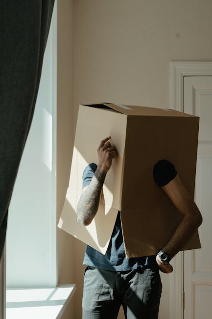 A man with a dark beard and curly hair, dressed in a dark blue T-shirt and matching trousers, is in a spacious room with white walls and wooden flooring. He is holding a cardboard box with red tape in