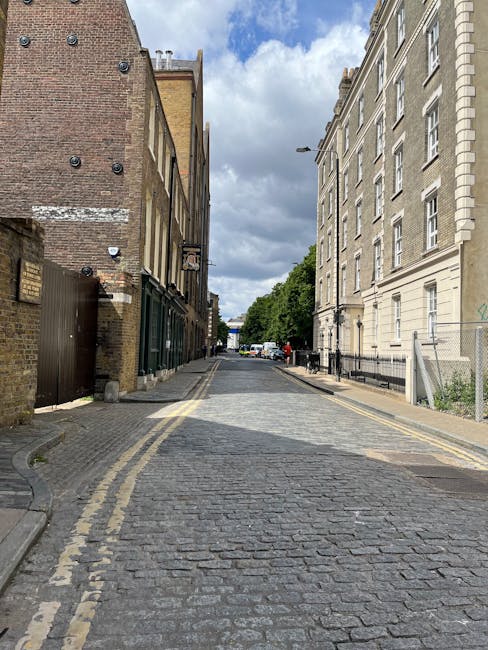 A narrow cobblestone street during daytime, lined with historical brick and stone buildings on both sides in Dalston, with some buildings featuring windows with white frames and decorative cornices. T