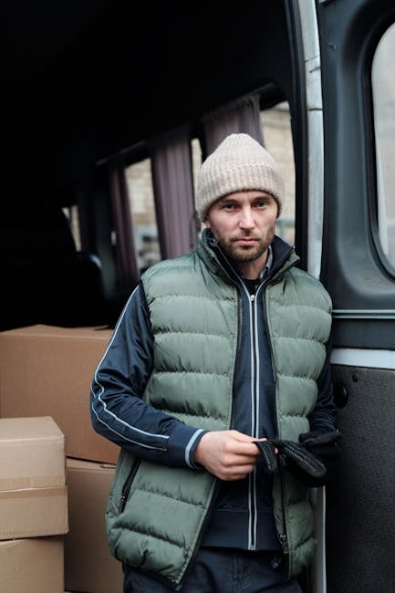 A man with a beard and wearing a red cap, plaid shirt, and a brown and beige work jacket stands inside a van with an open sliding door. Behind him, several cardboard boxes of various sizes, some taped